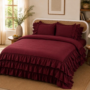 May include: A deep burgundy bedding set featuring a duvet cover with three tiers of ruffled edges and two matching ruffled pillow shams. The bedding is displayed on a bed frame with a light-colored woven headboard in a bright, neutral room.