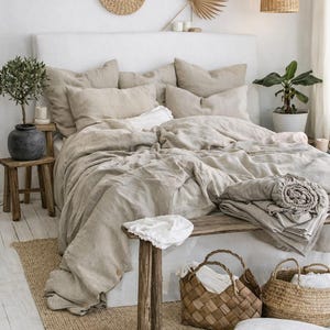 May include: A bedroom scene featuring a bed with beige linen bedding, pillows, and a headboard. Woven baskets, a small wooden stool, and a jute rug add to the room's natural aesthetic. Decorative wall hangings and a hanging wicker lamp complete the cozy space.