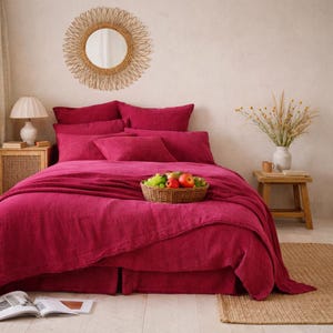 Può includere: Una camera da letto con un letto vestito con biancheria da letto rosa intenso, tra cui un copripiumino, cuscini e una gonna da letto. Un cesto di frutta intrecciato poggia sul letto. Uno specchio rotondo è appeso sopra il letto e un tavolino di legno con una lampada è a sinistra.