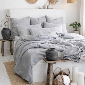 Pode incluir: Um quarto com uma cama com roupa de cama de linho cinza. O quarto apresenta uma paleta de cores neutras, com um tapete tecido, bancos de madeira e elementos decorativos como uma luminária pendente tecida e arte de parede.