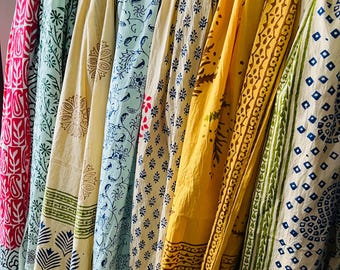 May include: A display of patterned fabrics in a range of colours, including pink, green, blue, yellow, and cream. The textiles feature diverse designs, such as floral and geometric patterns. These fabrics are likely used for home decor or clothing, hanging on a wooden rod.
