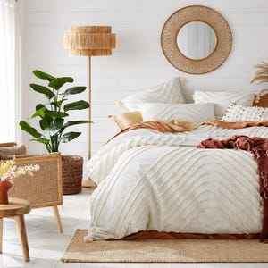 May include: A bedroom with a neutral colour scheme. The bed has a white duvet cover with textured, wavy patterns. A woven lamp and mirror enhance the bohemian style. A large potted plant and a small wooden table complete the decor.