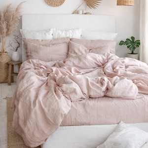 May include: A bedroom with a bed dressed in light pink linen bedding. The bed has a white headboard and is set against a white wall. Woven wall decorations and a woven pendant light add to the room's aesthetic. The room has a natural, calming feel.