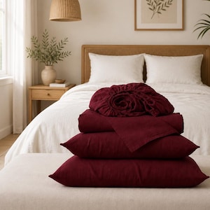 May include: A stack of deep burgundy linen bedding consisting of a fitted sheet, a flat sheet, and two rectangular pillowcases. The fabric has a visible woven texture and is folded neatly, resting on a light-colored upholstered bench.