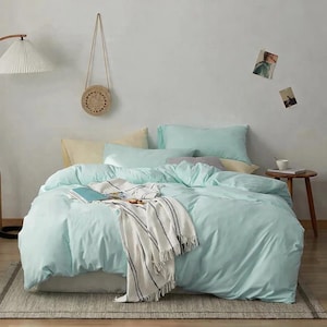 Puede incluir: Una cama con una funda nórdica azul claro y almohadas a juego. Una manta blanca con rayas azul oscuro está sobre la cama. Una pequeña mesita de noche de madera tiene una taza y un libro. Una bolsa tejida cuelga de la pared.