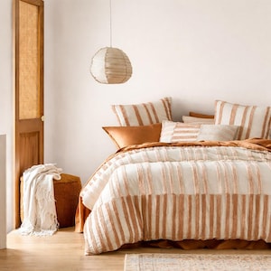 May include: A bed featuring a cream and rust-coloured striped duvet cover and matching pillows. The bed is styled with a rust-coloured bed skirt and a decorative cushion. A woven pendant light hangs above the bed, and a woven ottoman and blanket are in the corner.