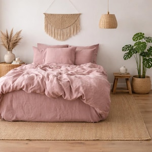 Puede incluir: Una escena de dormitorio con una cama con un edredón de lino rosa empolvado y almohadas a juego. Una alfombra de yute tejida se encuentra debajo de la cama. Los elementos decorativos incluyen un tapiz de macramé, una lámpara colgante de mimbre y una planta en maceta.