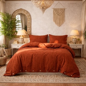 以下が含まれることがあります： ベッドに置かれた、錆色のベッドカバーと枕のセット。寝具は質感があり、ベッドの足元でボタン留めされています。お揃いの枕が2つ見えます。部屋には装飾的な鏡、ランプ、シャンデリアがあります。