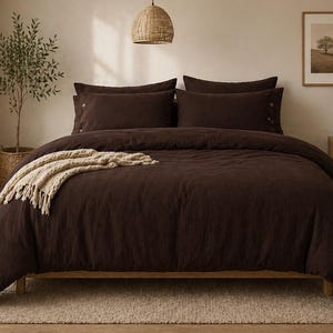 May include: A dark brown bed set with matching pillowcases and a duvet cover. A beige throw blanket with fringe is draped across the bed. The bedroom has a neutral color palette with a woven pendant light.