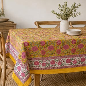 May include: A rectangular tablecloth featuring a vibrant mustard yellow base with a repeating pattern of pink floral blooms and green leaves. The border displays a distinct pink and white floral motif, adding a decorative edge to the fabric.