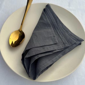 Puede incluir: Una cuchara dorada descansa sobre un plato color crema junto a una pila de servilletas de lino grises dobladas. La cuchara tiene una superficie reflectante. Las servilletas están cuidadosamente dispuestas, lo que sugiere un entorno de comedor.