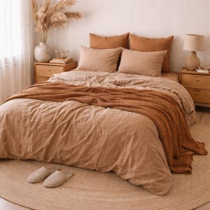 Op de afbeelding: Een slaapkamer met een bed met beige beddengoed en een roestkleurige deken. Twee houten nachtkastjes met lampen staan aan weerszijden van het bed. Onder het bed ligt een rond jute vloerkleed en een vaas met gedroogde planten.