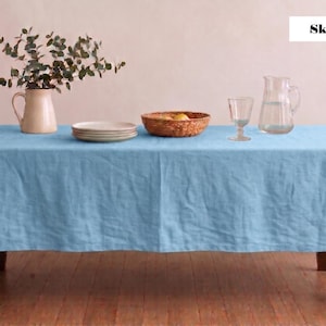 Puede incluir: Un mantel de lino azul cielo cubre una mesa de madera, con una jarra crema, platos, una cesta de mimbre, una copa y una jarra de cristal transparente. El texto "Sky Blue" está en la esquina superior derecha.