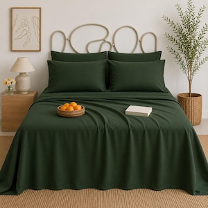 May include: A bed featuring dark forest green bedding, including a flat sheet and two matching pillowcases. A small woven basket containing several oranges sits on the bed, alongside a rectangular white book or box placed on the fabric.