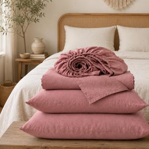 May include: A stack of dusty rose bedding consisting of two rectangular pillows and a folded flat sheet, topped with a gathered fitted sheet. The fabric has a visible woven texture, and the items are arranged neatly on a wooden bench.
