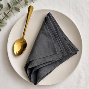 May include: A folded charcoal gray linen napkin rests on a white plate, next to a gold spoon. The spoon has a long handle and a rounded bowl. The napkin is neatly folded into a triangle shape.