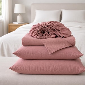 May include: A set of rose-pink bed linen, comprising a fitted sheet, a flat sheet, and two pillowcases. The bedding is stacked on a neutral surface, with a bed and lamp visible in the background. The sheets are made from a soft, textured material.