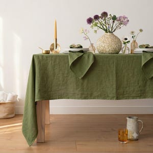 Puede incluir: Un mantel de lino verde oliva sólido cubriendo una mesa, con una superficie texturizada. La mesa está dispuesta con platos blancos, servilletas verde oliva dobladas, un candelabro dorado y un jarrón decorativo lleno de flores moradas y blancas.