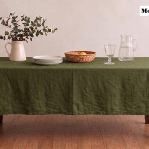 Puede incluir: Un mantel de lino verde musgo cubre una mesa de madera. Una jarra blanca con vegetación, una pila de platos, una cesta tejida con fruta, una copa de vino y una jarra de cristal están sobre la mesa. El texto "Moss Green" está en la esquina superior derecha.