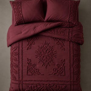 Peut inclure: Un ensemble de literie bordeaux profond composé d'une couette et de deux taies d'oreiller assorties. Le tissu présente une texture touffetée en relief formant des motifs floraux et géométriques complexes, incluant un médaillon central.