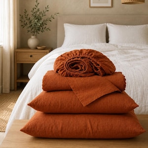 May include: A stack of burnt orange bedding items including a fitted sheet, a flat sheet, and two rectangular pillowcases. The fabric has a visible woven texture, and the items are neatly folded and layered on top of each other.
