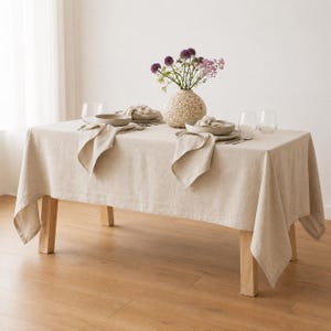 May include: A rectangular beige linen tablecloth draped over a wooden table. The table is set with two beige ceramic bowls, two folded beige linen napkins, four clear glass tumblers, and a textured beige vase containing purple flowers.