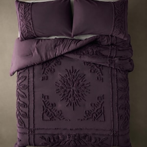 May include: A deep purple bedding set consisting of a comforter and two matching pillowcases. The fabric features a raised, textured floral and leaf pattern with a central sunburst motif, framed by decorative borders on the edges and pillows.