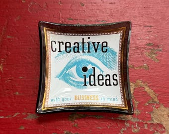 May include: A square glass trinket dish with curved edges, featuring a black border and a central graphic of a blue eye. The text reads "creative ideas" in black and "with your BUSINESS in mind" in smaller green and yellow lettering.