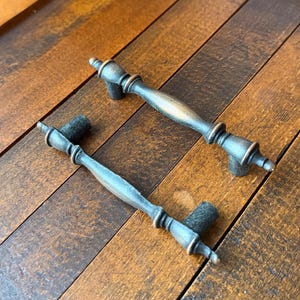 May include: Two metal drawer pulls featuring a dark, weathered bronze finish with subtle copper-toned highlights. Each handle has a central elongated body, decorative rings, and pointed finials at both ends, supported by two cylindrical mounting posts.