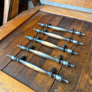 以下が含まれることがあります： 平行に並べられた5つの金属製引き出し用取っ手。各取っ手は、長く細くわずかに湾曲した中央バーと、両端の装飾的で丸みを帯びた段状のフィニアルが特徴です。金属は、使い古されたようなダークグレーとブロンズの仕上げになっています。