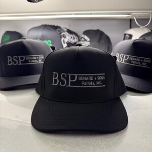 Pode incluir: Boné de beisebol preto com o texto bordado "BSP BENARD + SONS Pallets, INC." em branco. Vários bonés semelhantes são visíveis no fundo, sugerindo um item promocional ou uniforme.