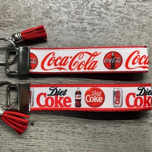 Puede incluir: Dos llaveros con borlas rojas. Un llavero presenta el logotipo de Coca-Cola en rojo sobre un fondo blanco. El otro llavero muestra el logotipo de Diet Coke en rojo sobre un fondo blanco. Ambos llaveros tienen herrajes plateados.