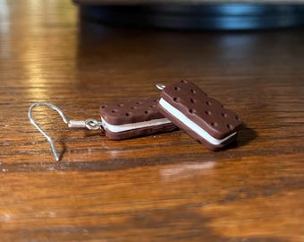 May include: A pair of novelty earrings shaped like ice cream sandwiches. The earrings feature a dark brown, rectangular cookie with a white cream filling. The earrings have silver-colored fish hook ear wires.