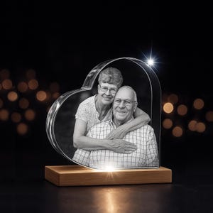 May include: A heart-shaped crystal photo with a black and white image of a smiling couple. The crystal sits on a rectangular wooden base, illuminated from below. The background is dark with bokeh lights.