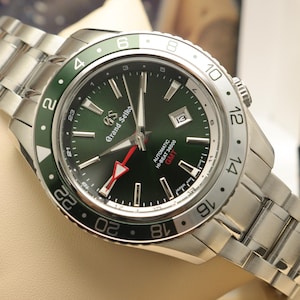 May include: A Grand Seiko automatic watch with a dark green dial, silver-tone hands and indices, and a date window at the three o'clock position. The watch features a stainless steel bracelet and a green and silver 24-hour GMT rotating bezel.