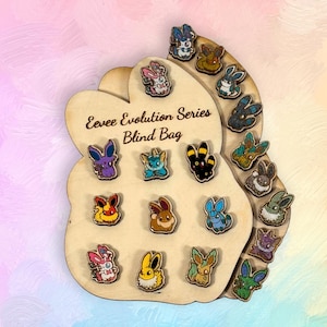 Könnte beinhalten: Holzdisplay mit einer Sammlung farbenfroher Hasen-Anstecknadeln aus der "Eevee Evolution Series Blind Bag". Die Pins im Cartoon-Stil sind auf einem Holzzuschnitt mit dem Serientitel angeordnet. Der Hintergrund ist eine weiche Mischung aus Rosa und Blau.