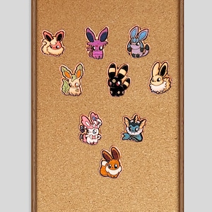 May include: A corkboard with a wooden frame displays nine colorful cartoon character pins. The pins feature various designs with bunny-like ears and different color schemes, including pink, purple, blue, green, black, and orange.
