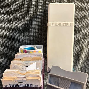 May include: A vintage View-Master set, including a white storage case, a viewer, and a purple reel holder filled with reels. The case and viewer are light gray. The reels have the text "ONE VIEW-MASTER REEL".