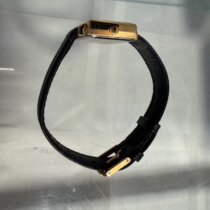 May include: A gold-toned rectangular wristwatch with a black leather band. The watch has a simple, elegant design with a gold buckle. The band is curved, and the watch is displayed against a neutral background.