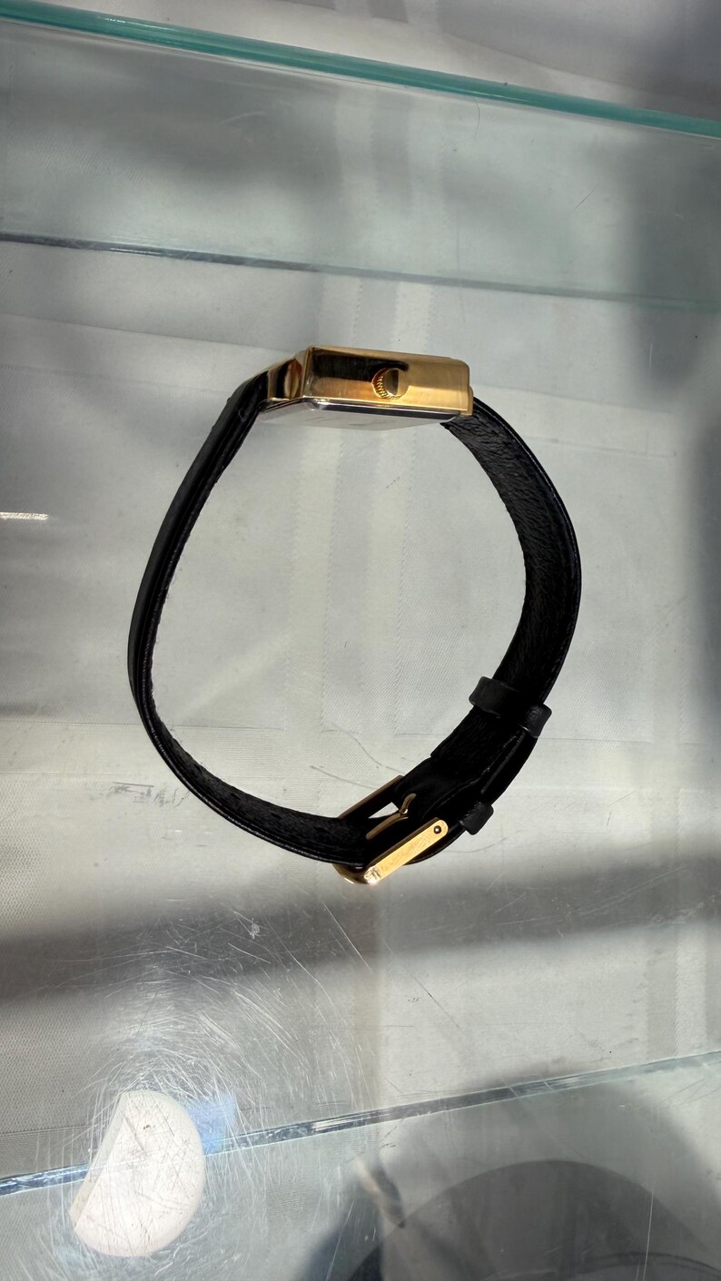 May include: A gold-toned rectangular wristwatch with a black leather band. The watch has a simple, elegant design with a gold buckle. The band is curved, and the watch is displayed against a neutral background.