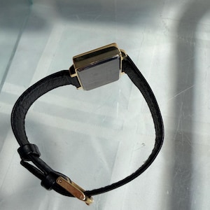May include: A rectangular wristwatch with a black leather band and a gold-colored case. The watch face is dark. The brand name "FLAGTIME" is visible on the box. The watch has a classic, minimalist design.