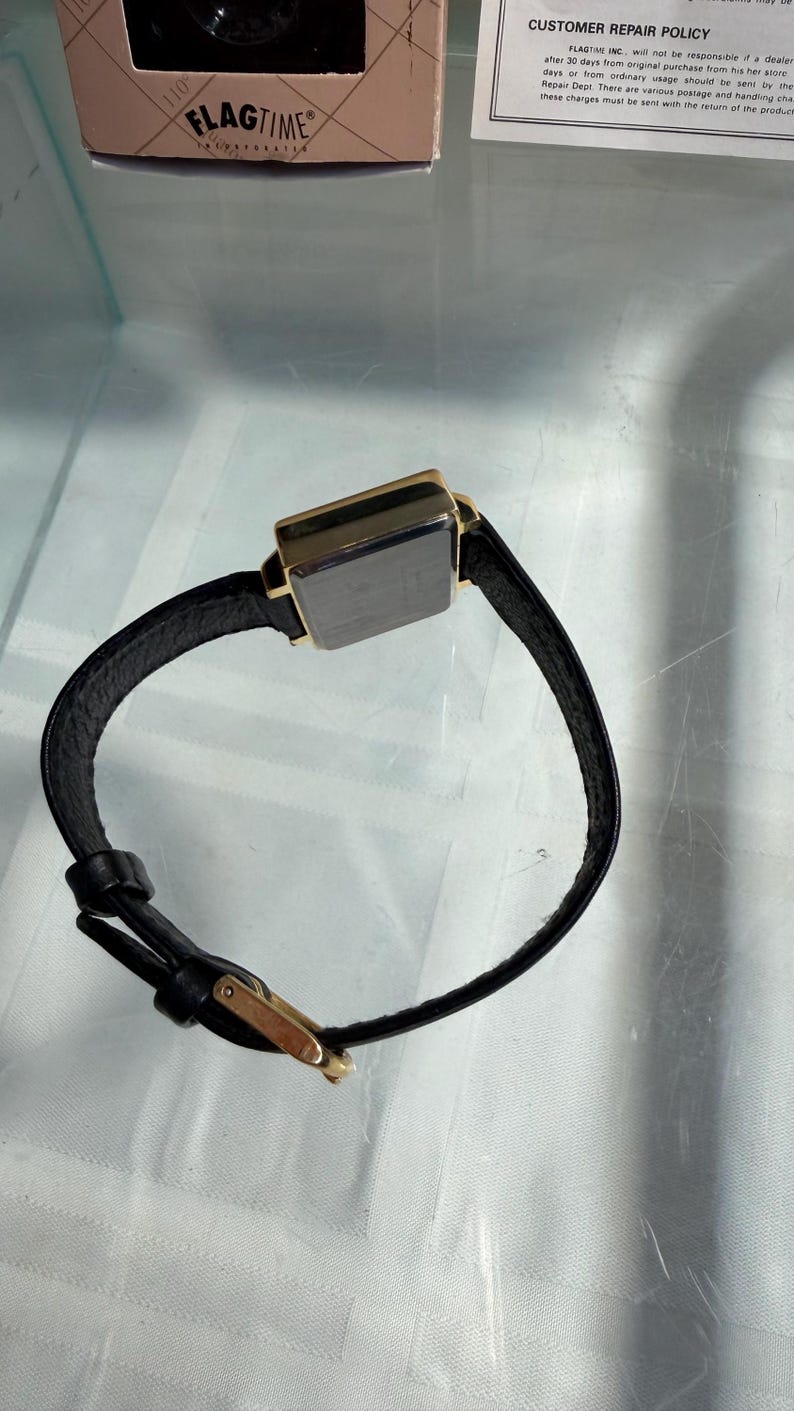 May include: A rectangular wristwatch with a black leather band and a gold-colored case. The watch face is dark. The brand name "FLAGTIME" is visible on the box. The watch has a classic, minimalist design.