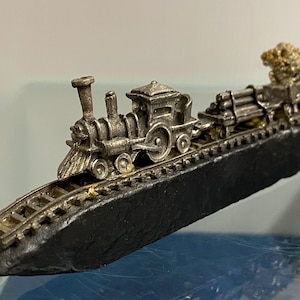 May include: A miniature silver-coloured train set with a locomotive, cargo cars, and a coal car. The train is on a black track. The train cars are loaded with logs and a pile of gold-coloured material.