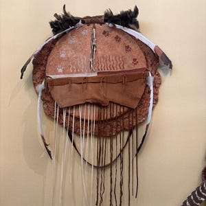 May include: A handcrafted, circular wall hanging with a rustic aesthetic. It features a central, light brown section adorned with paw prints and vertical lines. Brown leather pouches and hanging strings are attached, with feathers and fur accents.