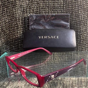 May include: Red Versace spectacles with a matching case. The glasses have a rectangular frame with a decorative design on the temples. The case is black with the Versace logo in gold lettering.