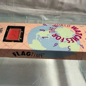 May include: A vintage Flagtime watch in its original box. The watch has a red face and gold-colored frame. The box is peach-colored with a world map graphic and the text "The World Watches For". The brand name "FLAGTIME" is printed on the box.