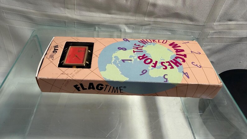 May include: A vintage Flagtime watch in its original box. The watch has a red face and gold-colored frame. The box is peach-colored with a world map graphic and the text "The World Watches For". The brand name "FLAGTIME" is printed on the box.
