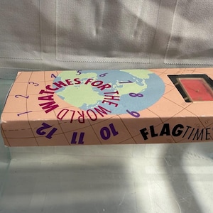 May include: A rectangular box with the words "WATCHES FOR THE WORLD" and "FLAGTIME" printed on it. The box has a peach background with a world map graphic and numbers around the edge. A watch is visible through a window.