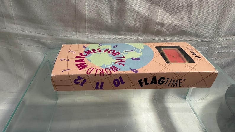 May include: A rectangular box with the words "WATCHES FOR THE WORLD" and "FLAGTIME" printed on it. The box has a peach background with a world map graphic and numbers around the edge. A watch is visible through a window.