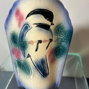 May include: Ceramic wall pocket vase with a bird design. The vase has a cream-colored center with a blue border and a bird illustration in black and blue. Pink and green accents complete the design.